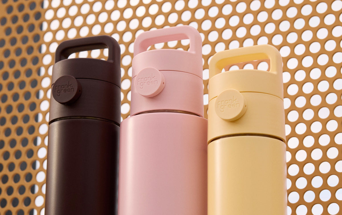 Three Frank Green insulated water bottles in dark brown, light pink, and pale yellow, standing side-by-side on a gray surface in front of a warm orange perforated metal screen.