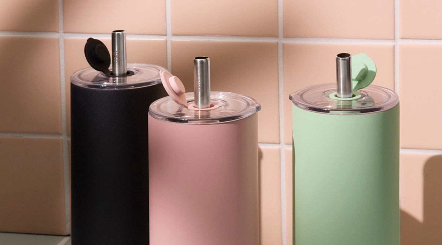 Three insulated tumblers in matte black, blush pink, and sage green with clear lids and metal straws, arranged against a beige tiled background.