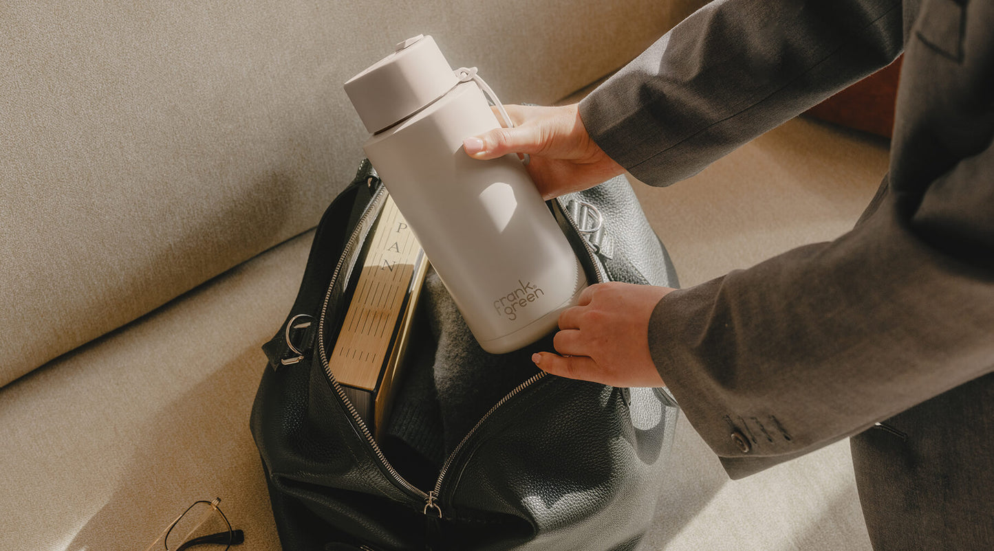 A person in a gray suit packs a light gray reusable bottle into a black leather bag next to books on a beige sofa.