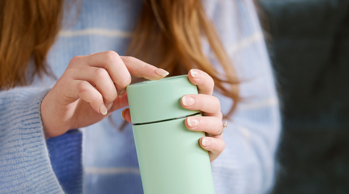 A close-up of a person holding a matte mint green travel tumbler with a matching lid, shown in a lifestyle setting with a cozy background.