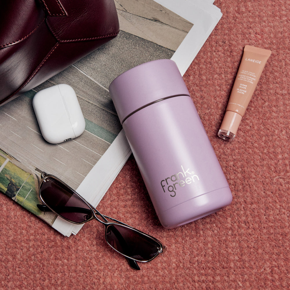 A matte lilac purple Frank Green vacuum insulated travel tumbler bottle, styled in a flat-lay arrangement on a textured terracotta surface with sunglasses, a white earbud case, a makeup tube, and a brown leather bag.
