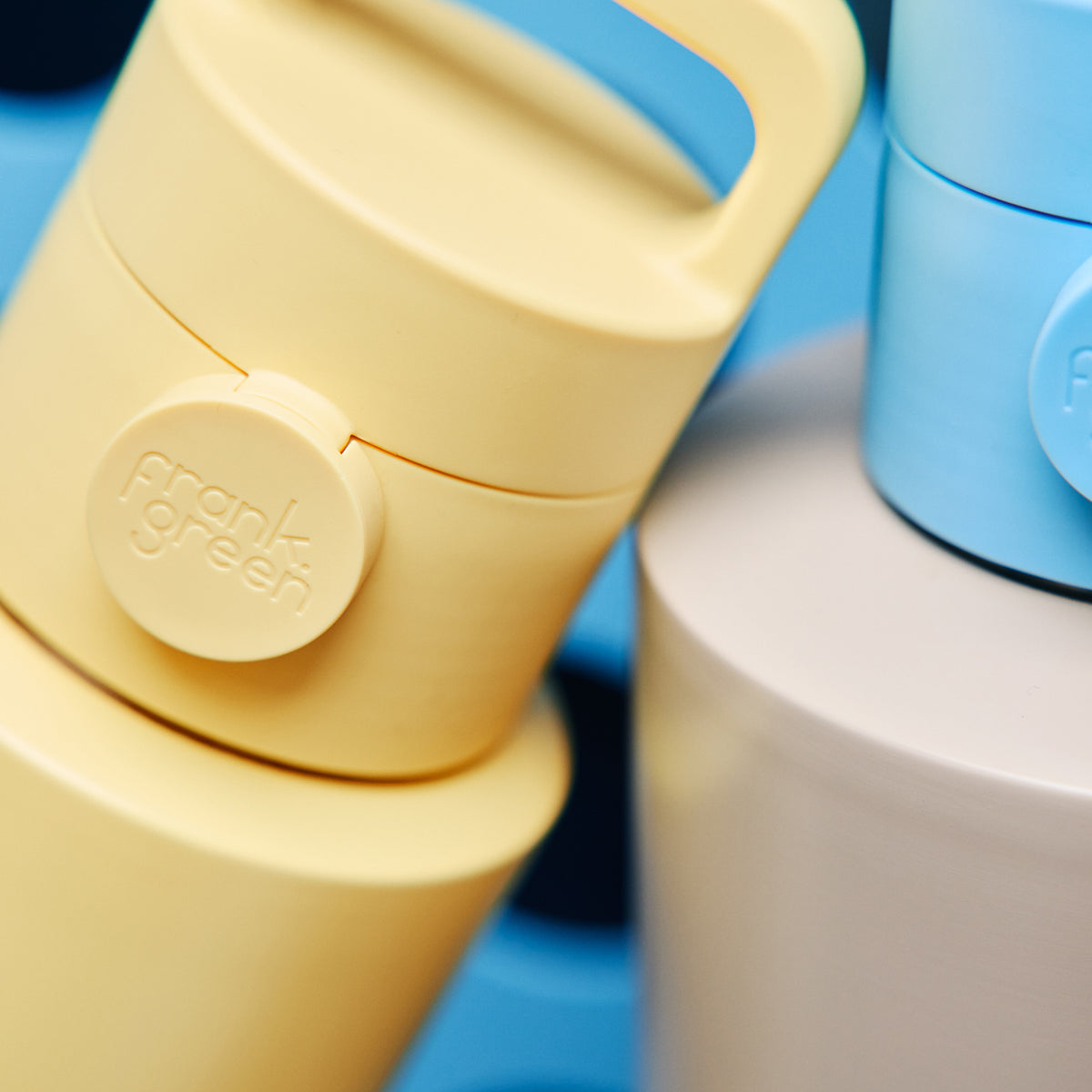 Close-up of a Frank Green travel tumbler with a pale yellow lid and handle, showing the embossed brand name on the side, with other tumblers partially visible in blue and white.