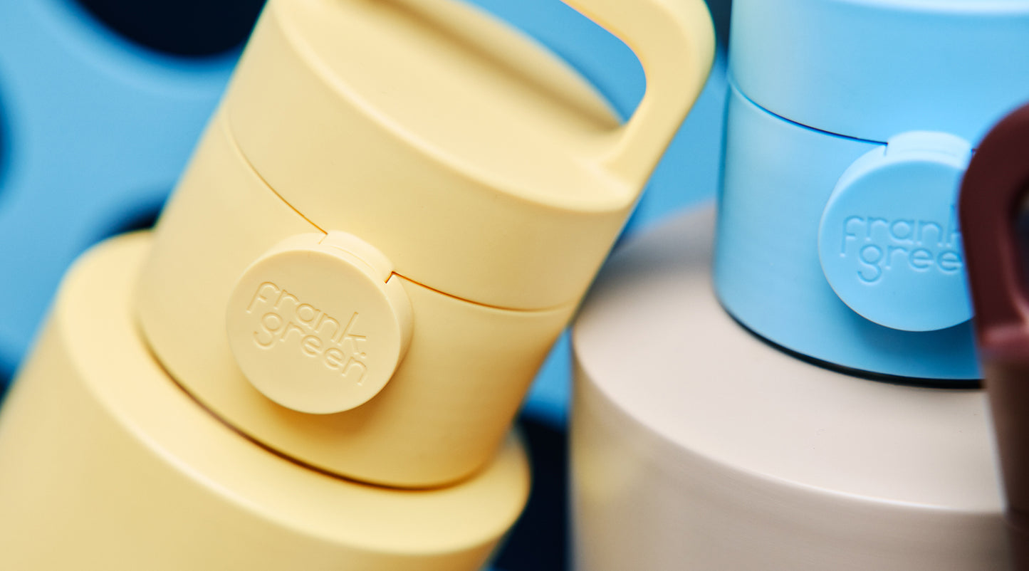 Close-up of a Frank Green reusable bottle lid in pale yellow, with part of a blue bottle and a white container visible in the background.