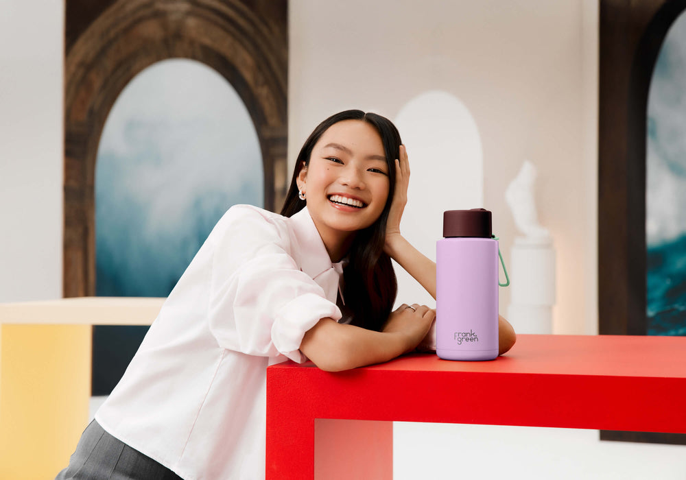 A woman in a white shirt leans on a red table next to a lavender insulated water bottle with a dark brown lid. The background features modern architectural elements and colorful geometric shapes.