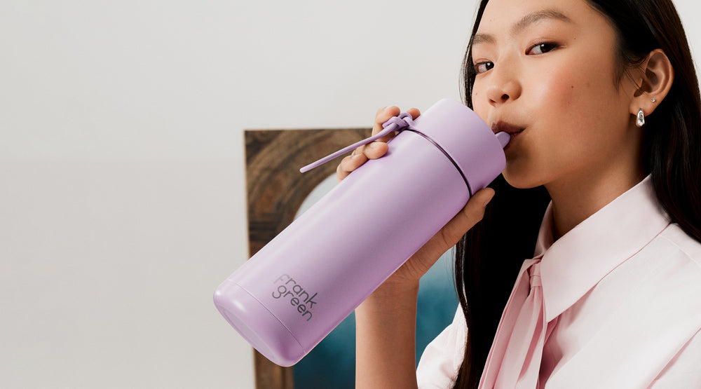 A woman in a light pink shirt drinking from a lavender purple insulated water bottle with a matching straw. The bottle features the 'park green' brand logo near the bottom.