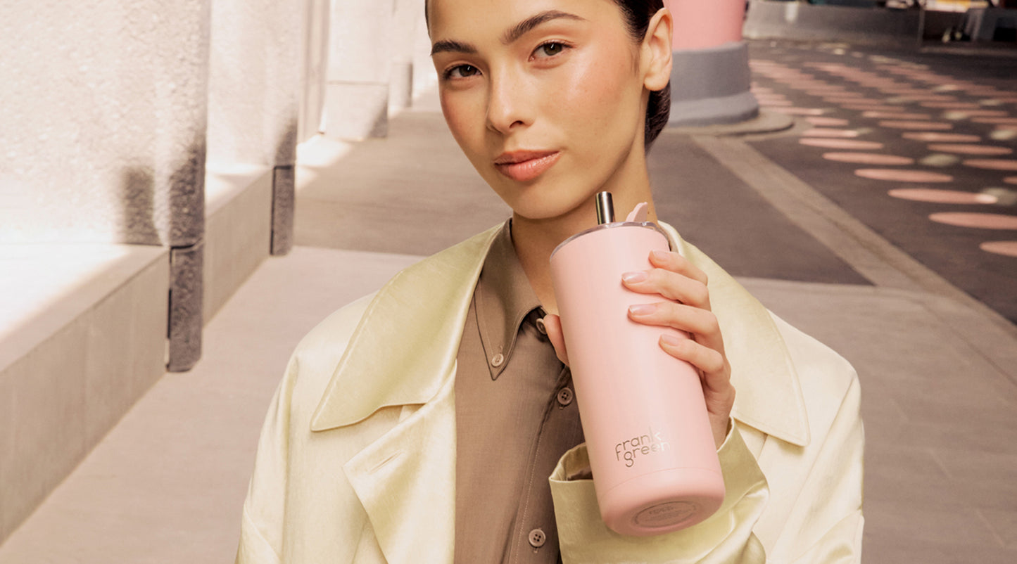 A woman in a cream trench coat holds a light pink frank green insulated tumbler with a silver lid and straw, set against a softly blurred urban walkway background.