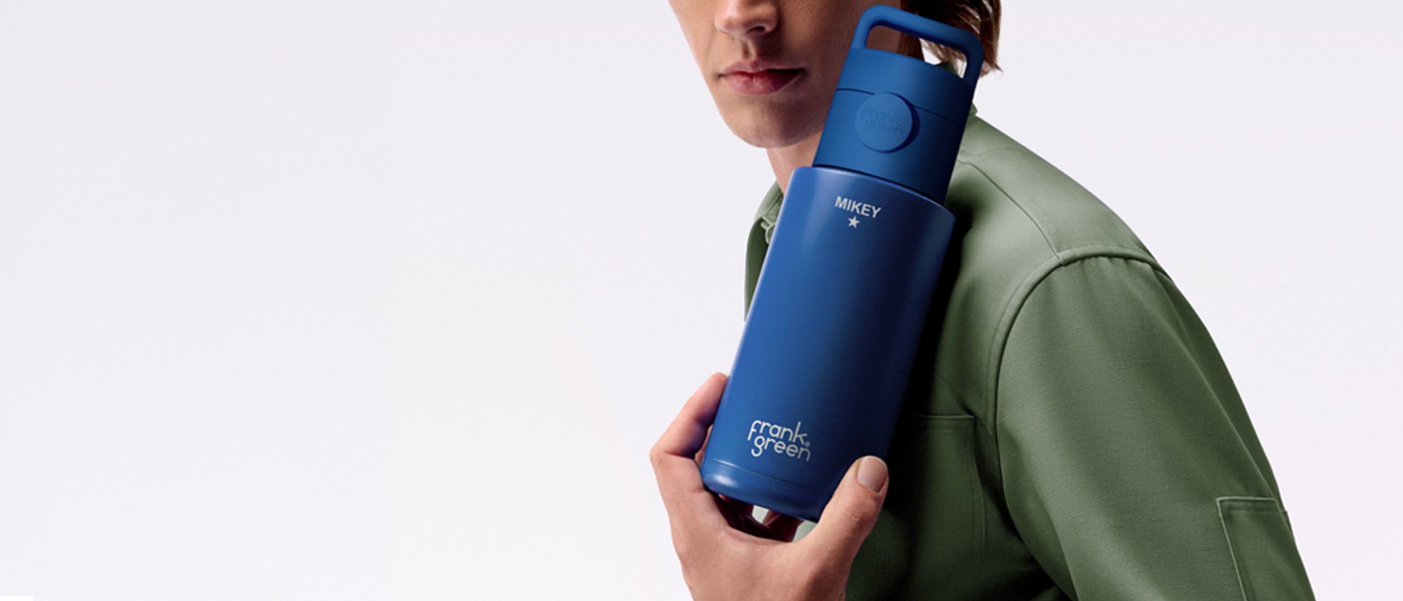 A man in a green jacket holds a blue insulated travel mug with a matching blue lid against a clean light gray studio background.