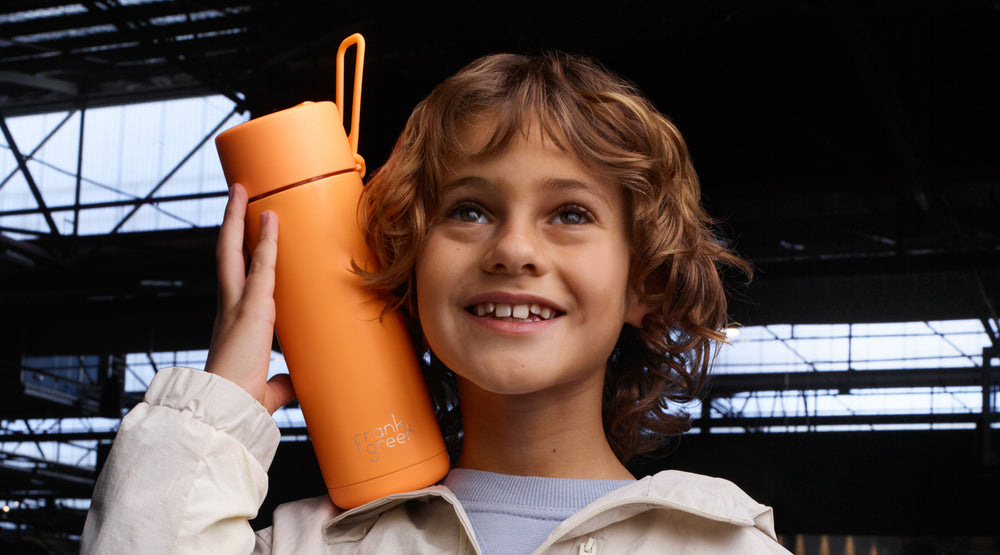 A smiling young boy with curly light brown hair holds an orange insulated water bottle against his shoulder. The bottle has a matching orange lid with a carrying loop and a small white logo near the bottom.