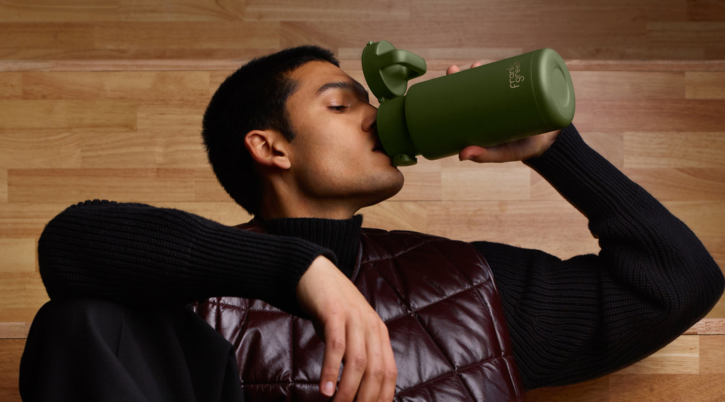 A man drinking from an olive green insulated water bottle with a flip-top lid, wearing a black ribbed sweater and a dark brown quilted vest against a light wood-paneled background.