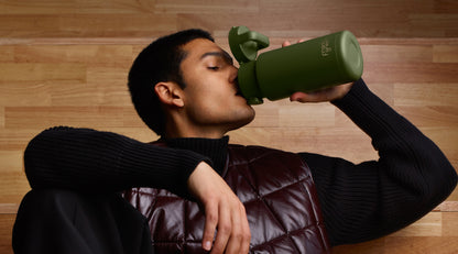 A man drinking from an olive green insulated water bottle with a flip-top lid, wearing a black ribbed sweater and a dark brown quilted vest against a light wood-paneled background.