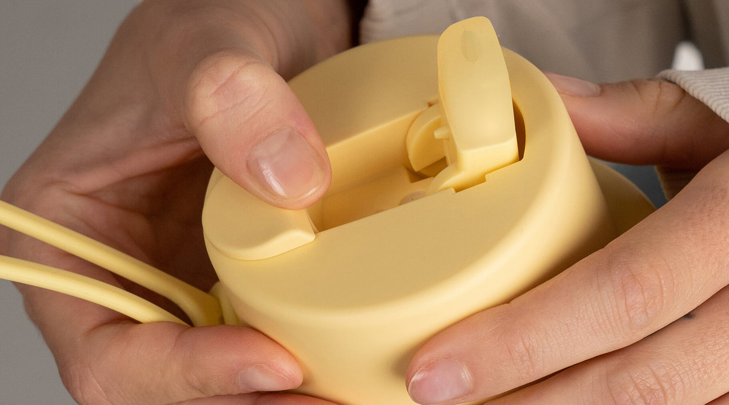 Close-up of a person's hands holding a pale yellow reusable water bottle with a flip-top lid, demonstrating how to open it.