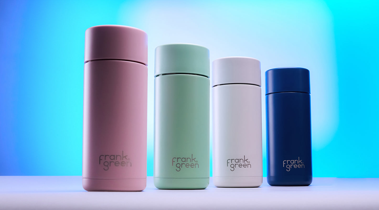 Four Frank Green vacuum insulated tumblers in pink, mint green, white, and navy blue, lined up on a white surface against a blue gradient background.