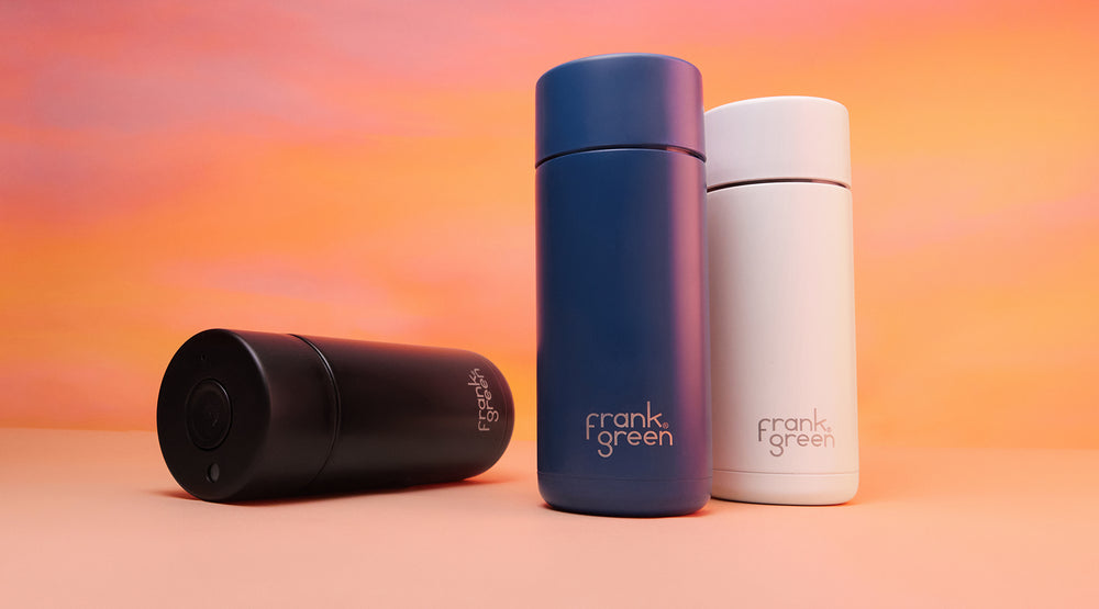 Three insulated travel tumblers by Frank Green are displayed on a peach-colored surface against a warm orange gradient background. The set includes one black tumbler lying on its side, one dark blue tumbler standing upright, and one white tumbler standing upright.