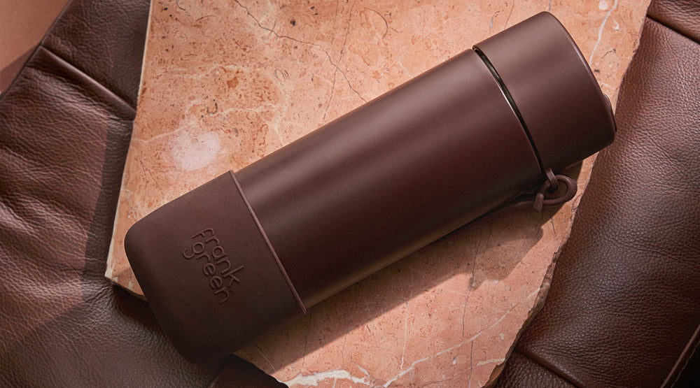 A dark brown vacuum-insulated travel tumbler with a matching lid and a small loop handle, resting on a textured brown leather surface with a warm stone background.