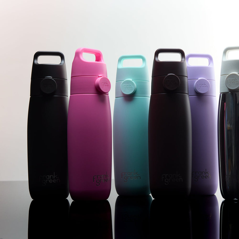 Six reusable insulated bottles in different colors lined up on a reflective black surface against a light gray background. The bottles are black, magenta, mint green, dark brown, lavender, and silver.