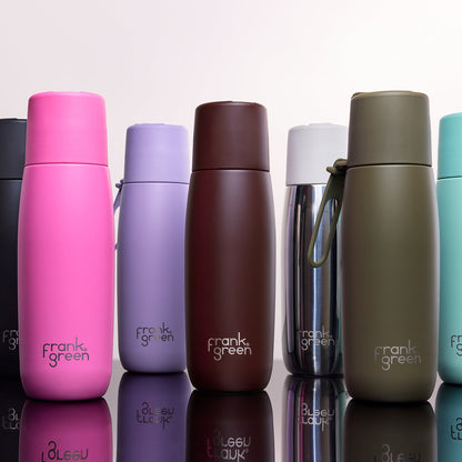 A lineup of Frank Green stainless steel water bottles in various colors, including pink, purple, brown, silver, and olive green, arranged on a reflective white surface against a light gray background.