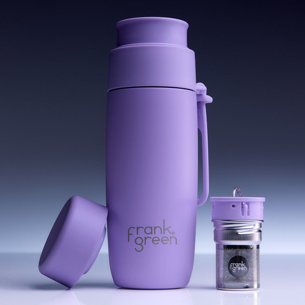 A purple Frank Green vacuum-insulated bottle with its lid off, shown next to a small matching purple and silver tea infuser on a clean studio background.