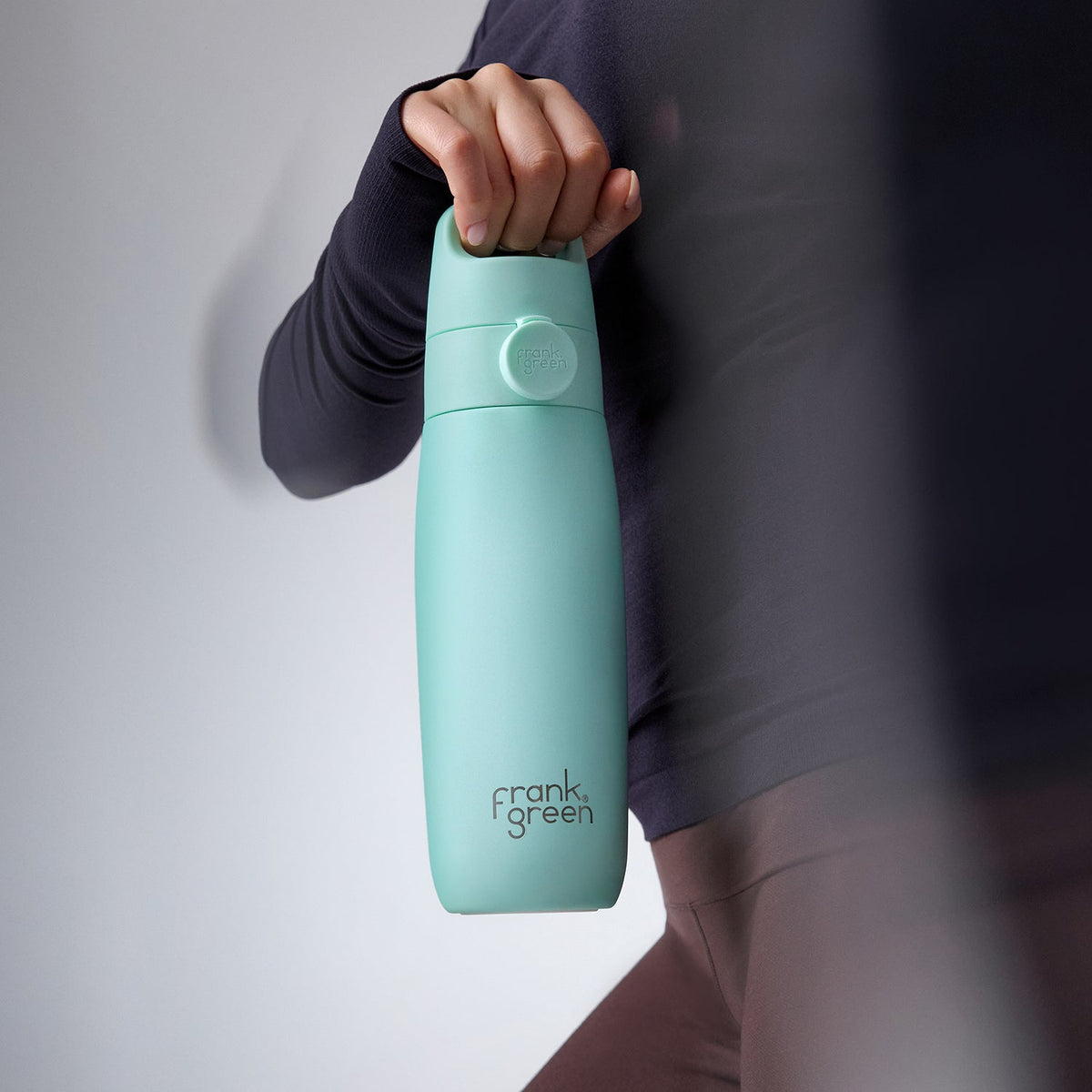 A person in a dark long-sleeve top holds a mint green Frank Green water bottle against a light gray studio background.