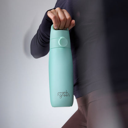 A person in a dark long-sleeve top holds a mint green Frank Green water bottle against a light gray studio background.