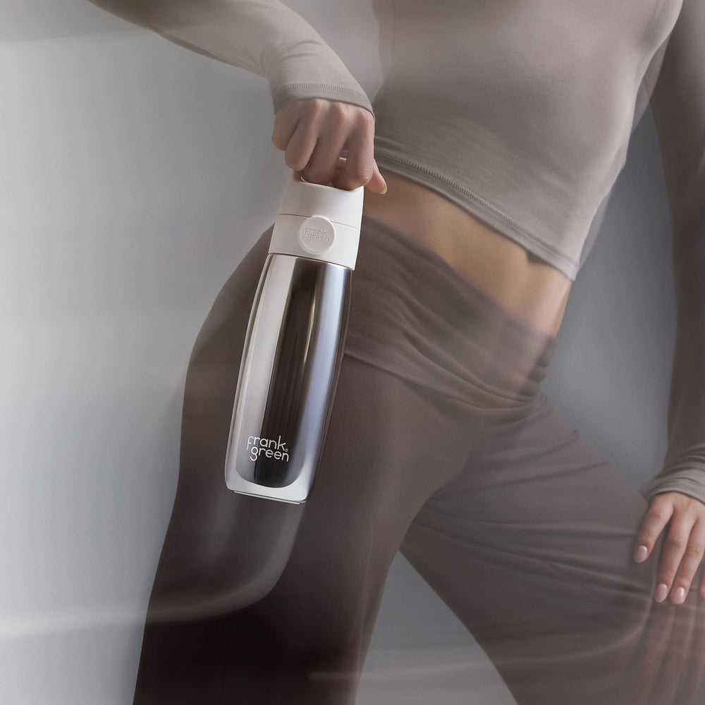 A person in taupe activewear holds a sleek, metallic silver and white Frank Green water bottle against a minimalist gray background.