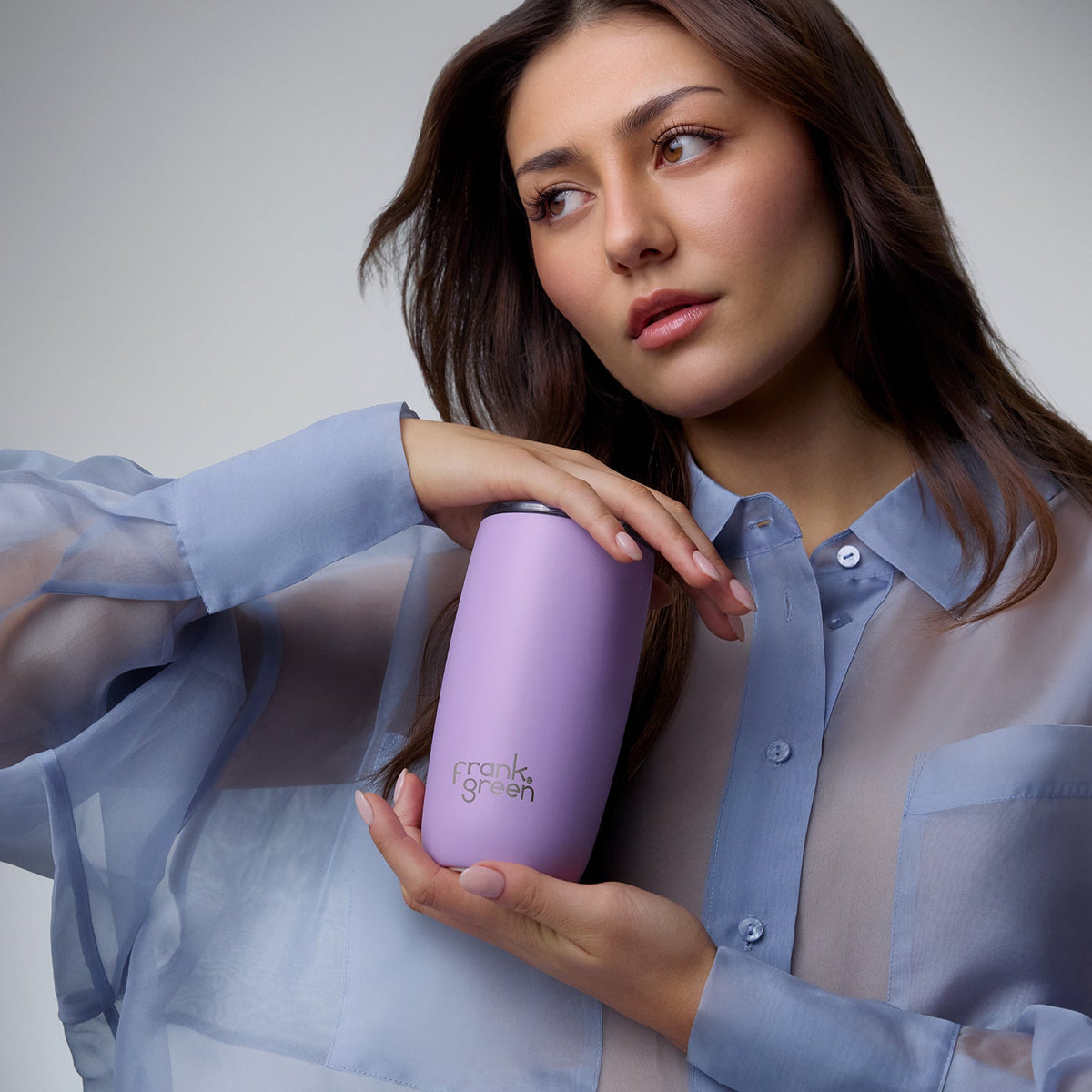 A woman in a sheer light blue button-up shirt holds a lavender purple insulated tumbler with the brand name 'frank's garden' printed on it, against a neutral gray studio background.