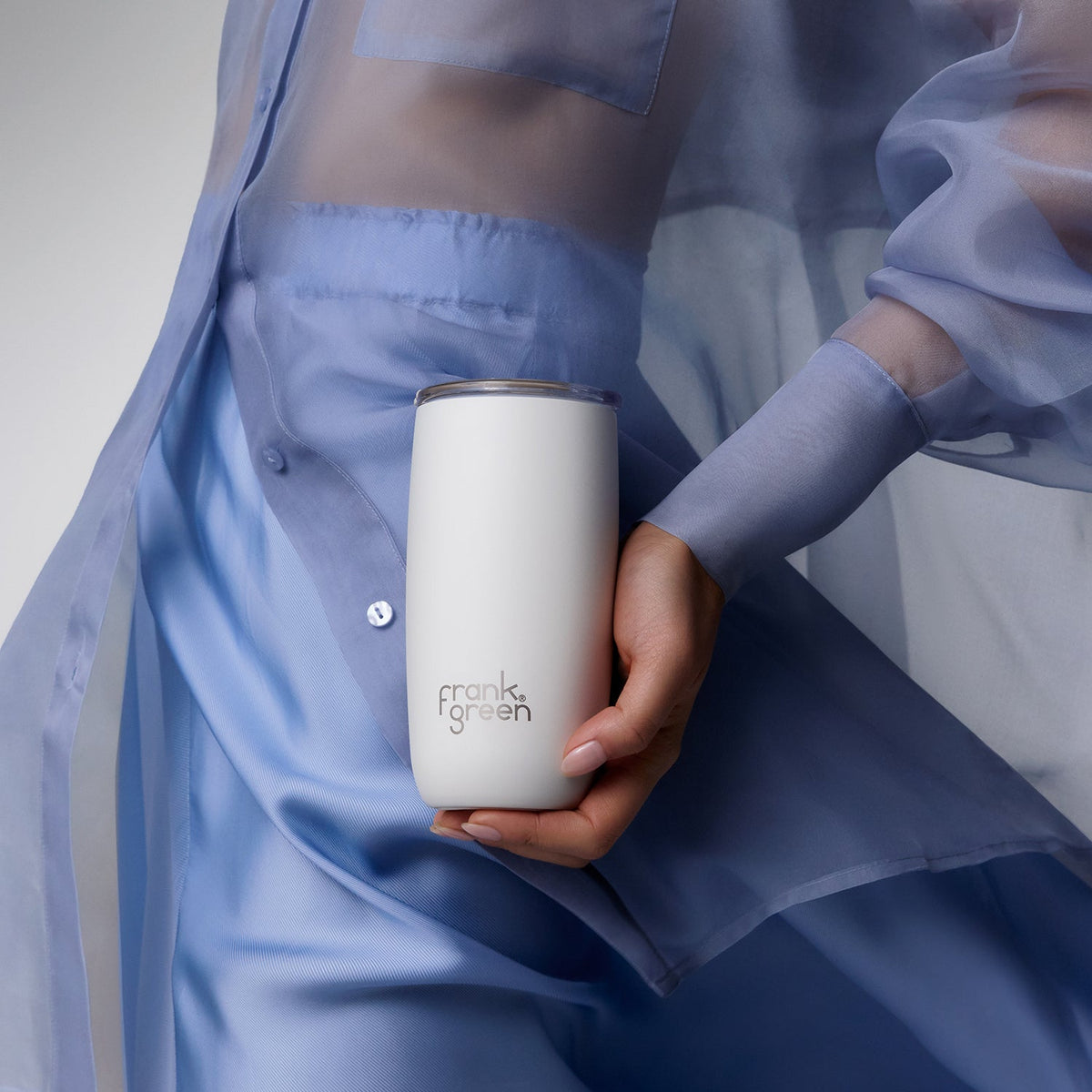 A person in a sheer light blue dress holds a white insulated tumbler with a silver rim. The tumbler has the 'frank green' logo printed on the front.