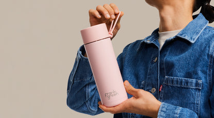 A person in a blue denim shirt holds a light pink insulated water bottle with a matching pink loop handle. The bottle has the 'Frank' brand name printed near the bottom.