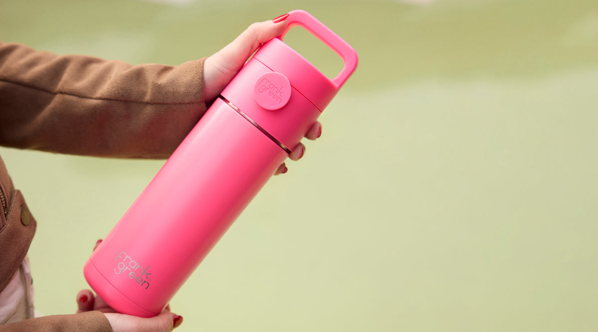 A person holds a bright pink vacuum-insulated water bottle with a matching carrying loop lid against a soft, out-of-focus pale green background.