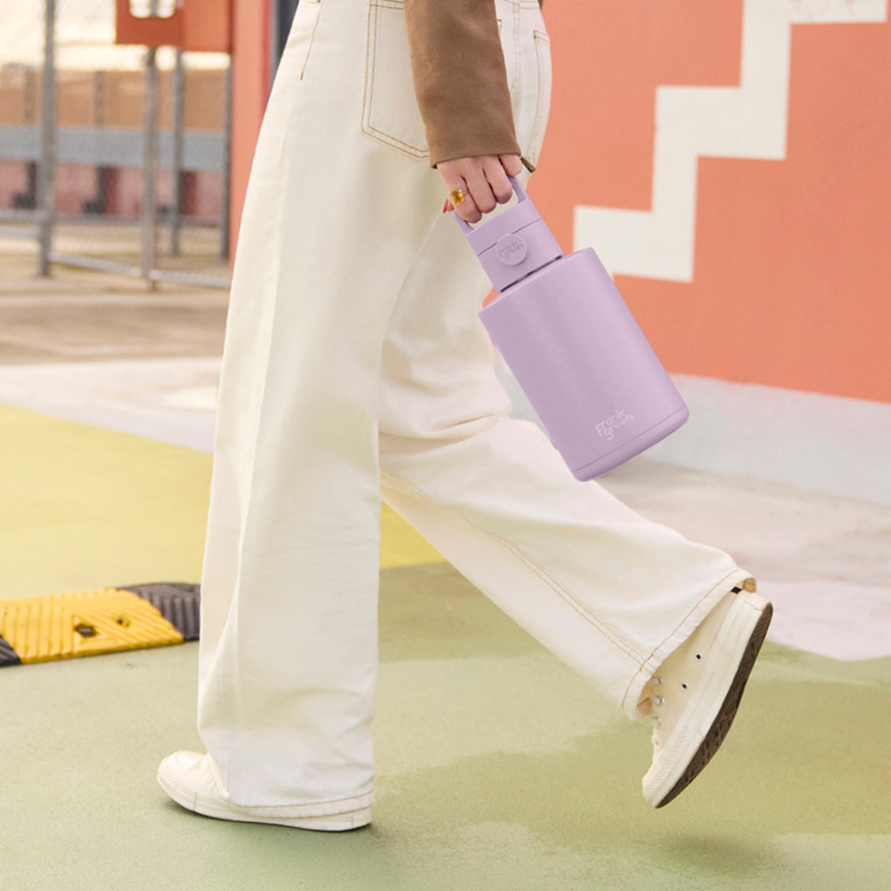 A person walking outdoors while holding a light purple reusable bottle. The person is wearing off-white wide-leg pants, white sneakers, and a brown jacket.