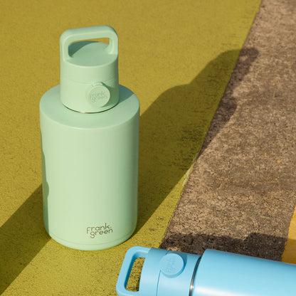 A mint green Frank Green insulated bottle with a matching lid and carry loop, photographed outdoors on a yellow surface beside a concrete curb. A second bottle in light blue is partially visible in the foreground.