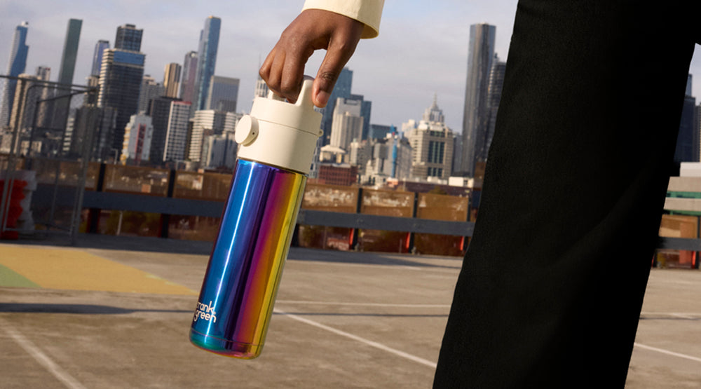 A person holding a metallic iridescent water bottle with a white lid, set against a blurred city skyline and rooftop background.