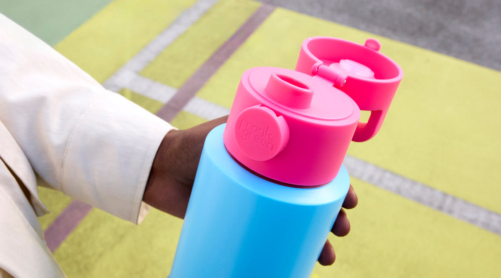 A close-up of a person holding a reusable water bottle with a bright blue body and a pink flip-top lid. The background shows a blurred outdoor court surface in yellow-green tones.