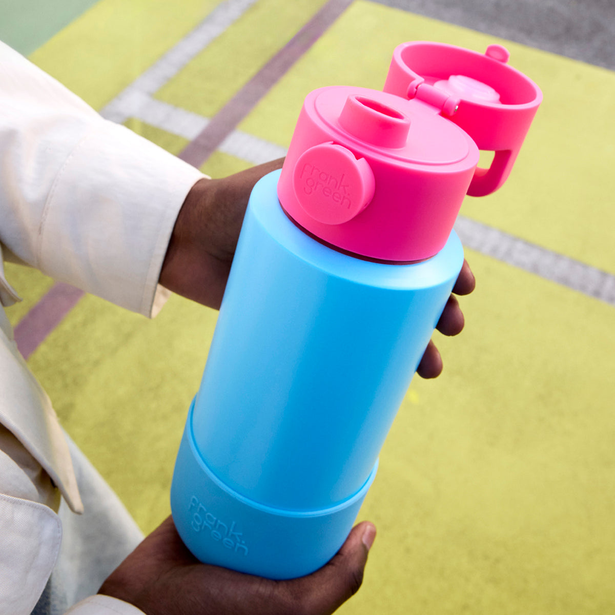 A person holding a reusable water bottle with a bright blue body and a pink flip-top lid. The bottle has a matching pink cap and a pink base.
