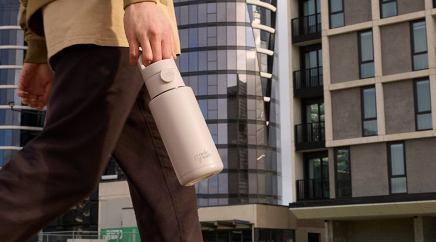 A person walking outdoors in an urban setting, carrying a matte light beige insulated water bottle. Modern glass and concrete buildings are visible in the background.