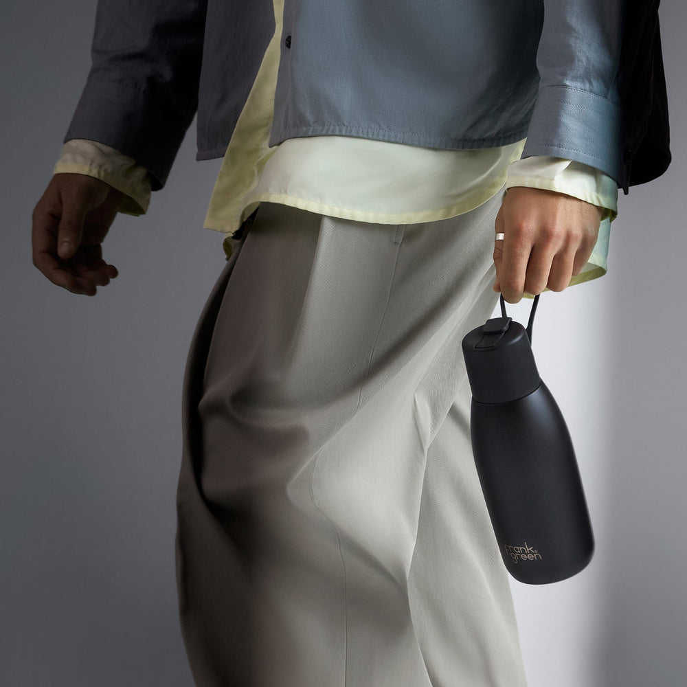 A person wearing a layered outfit with a gray jacket, cream shirt, and light gray trousers holds a matte black insulated bottle with a small gold logo. The background is a clean, neutral gray studio backdrop.