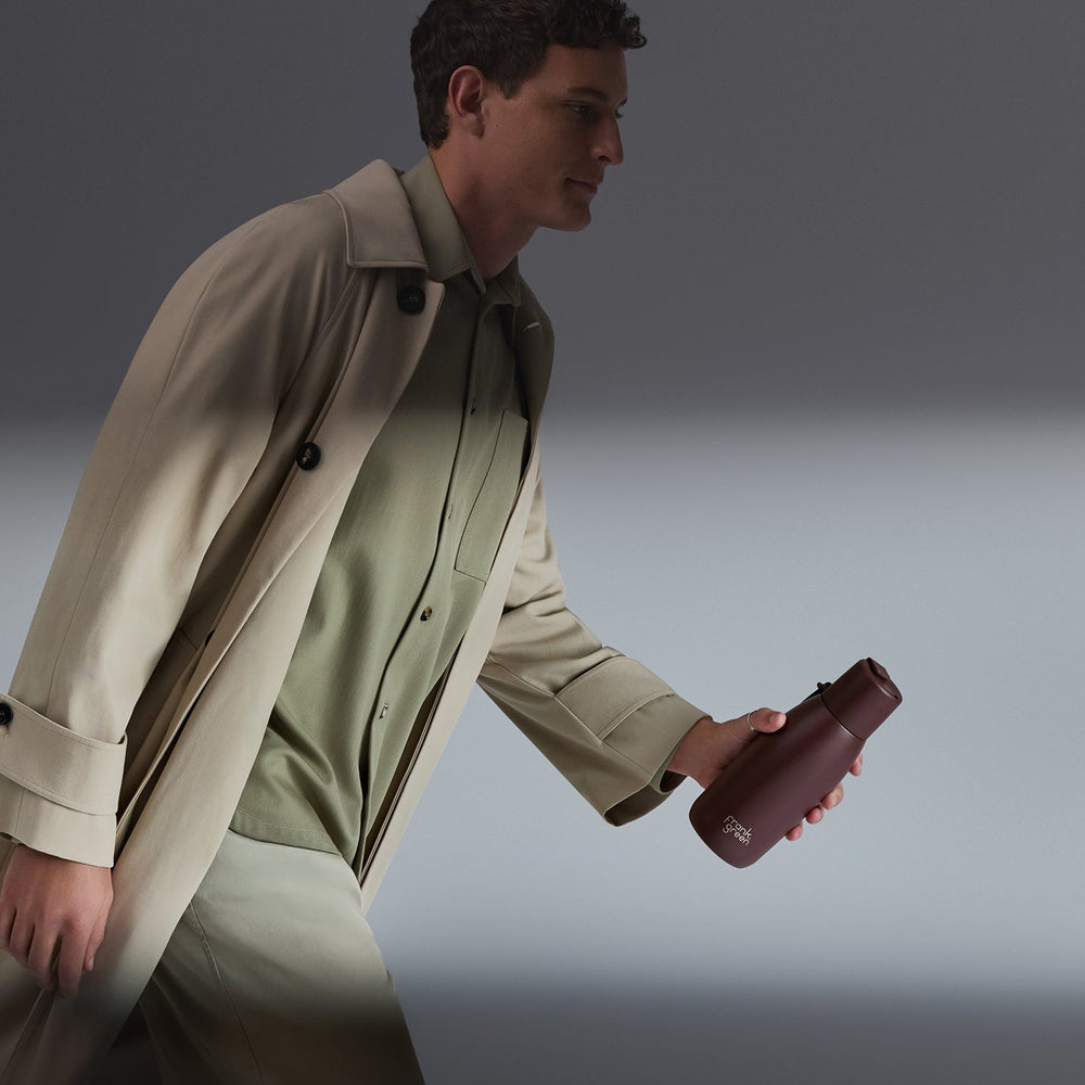 A man in a beige trench coat and light green shirt walks while holding a dark brown insulated water bottle with a visible POLYWOOD logo, set against a dark gray studio background.