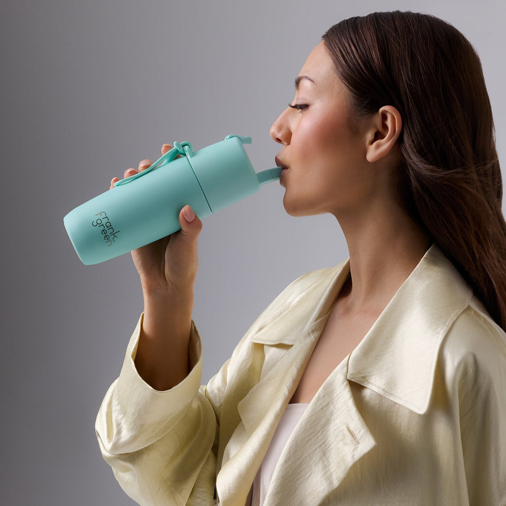 A woman holds up a small bottle with a sipper to her mouth and takes a drink.