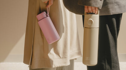 Two people holding insulated water bottles. One person in a beige coat holds a pink bottle, and the other in a gray coat holds a light beige bottle.