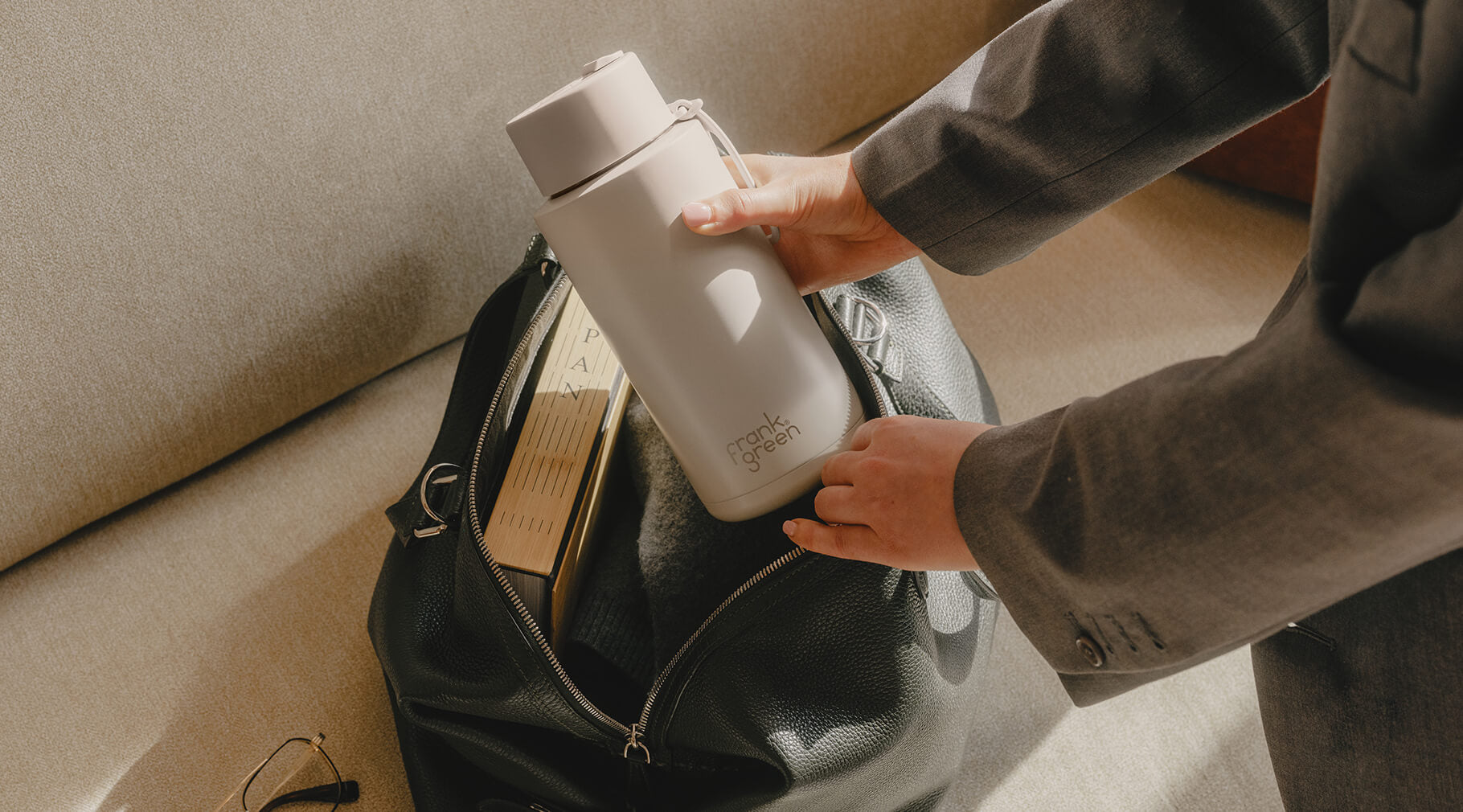 A person in a gray suit packs a light gray reusable bottle into a black leather bag next to books on a beige sofa.