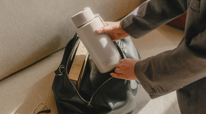 A person in a gray suit packs a light gray reusable bottle into a black leather bag next to books on a beige sofa.